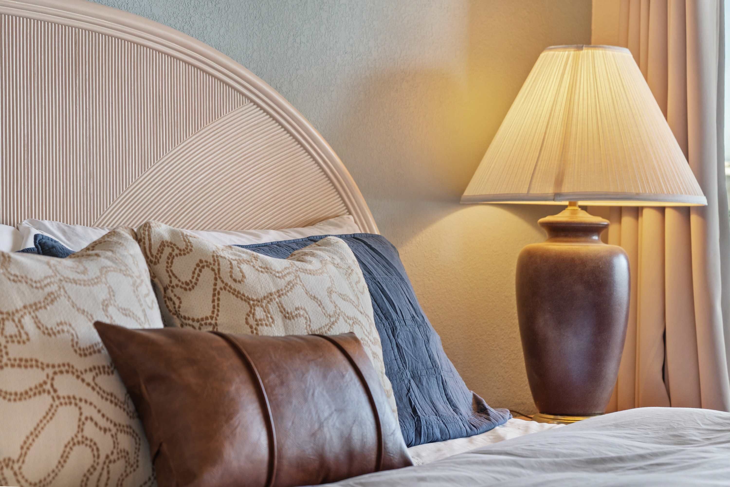 A neatly made bed with patterned pillows and a brown leather pillow, a round wooden headboard, and a tall floor lamp with a beige lampshade.
