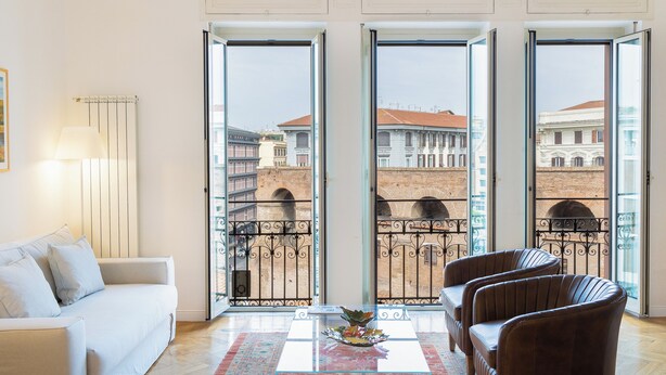 A room with a white sofa, two brown armchairs, a glass coffee table with a floral centerpiece, and a view of a cityscape through large glass doors. - Amazing Piazza Fiume (Roma)