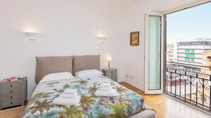 A bedroom with a bed, bedside tables, a nightstand, a window with a view of buildings, and a balcony with a railing. - Amazing Piazza Fiume (Roma)