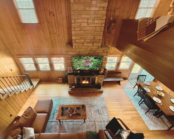 Una sala de estar acogedora con chimenea de piedra, paredes de madera, televisor, sofá y comedor.