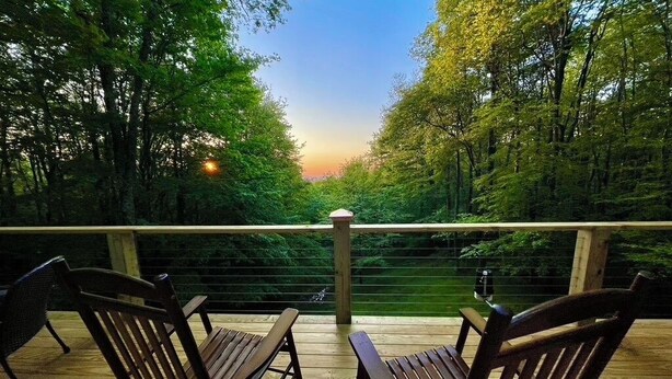 Una terraza de madera con dos sillas mirando hacia un área boscosa, una barandilla y una puesta de sol al fondo.