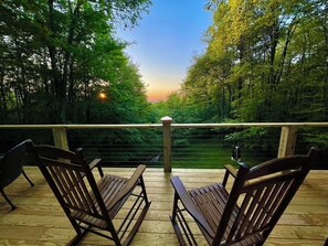Una terraza de madera con dos sillas mirando hacia un área boscosa, una barandilla y una puesta de sol al fondo.