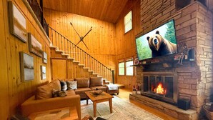 Interior acogedor de una cabaña de troncos con chimenea de piedra, un televisor mostrando un oso y una escalera con barandillas de madera.