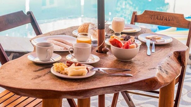 A wooden table with breakfast items including eggs, toast, and fruit, set against a poolside view. - Pousada Praia dos Corais (Paripueira)