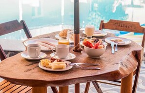 A wooden table with breakfast items including eggs, toast, and fruit, set against a poolside view. - Pousada Praia dos Corais (Paripueira)