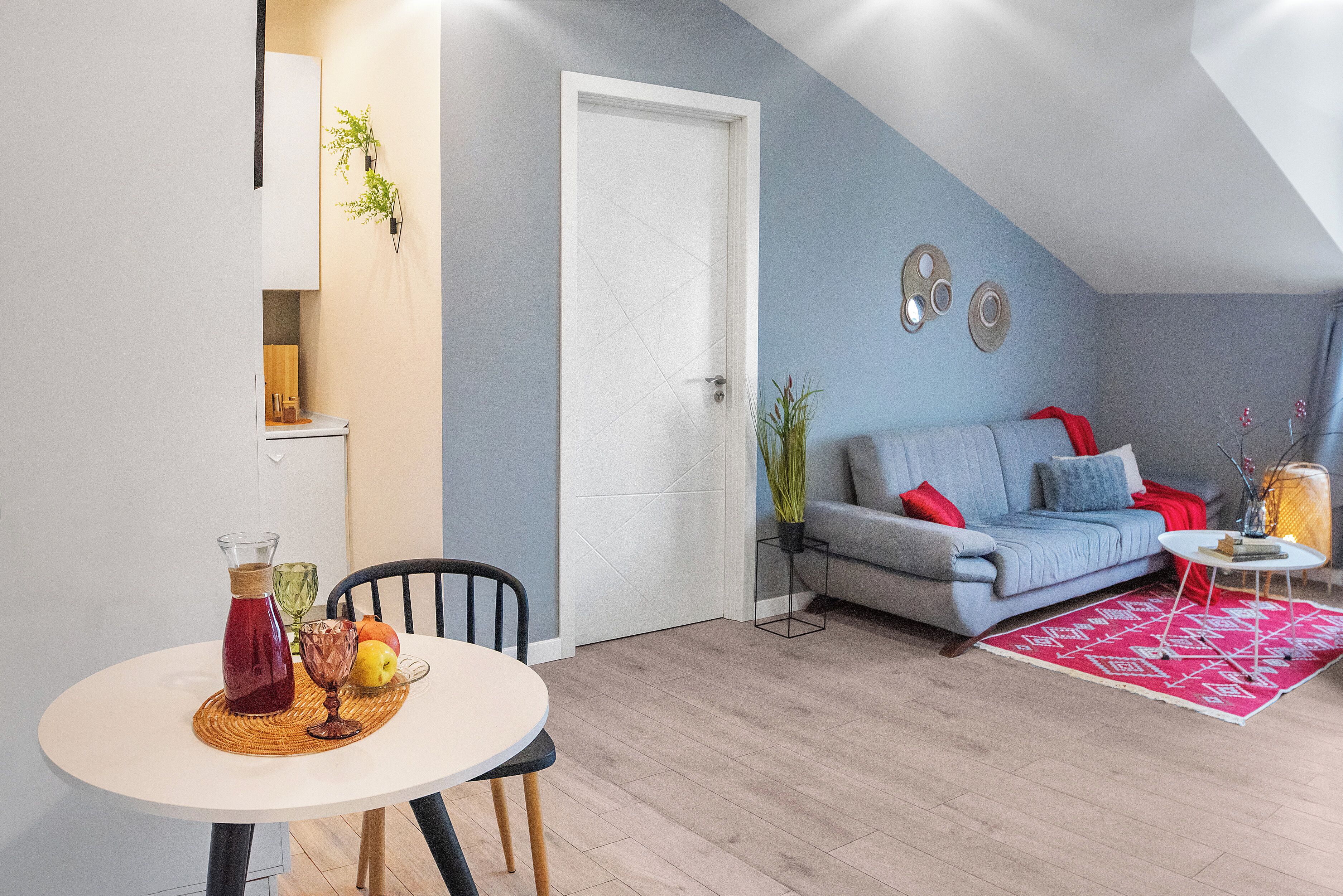 A modern living room with a grey sofa, a dining table with a pitcher and fruit, and a red patterned rug.
