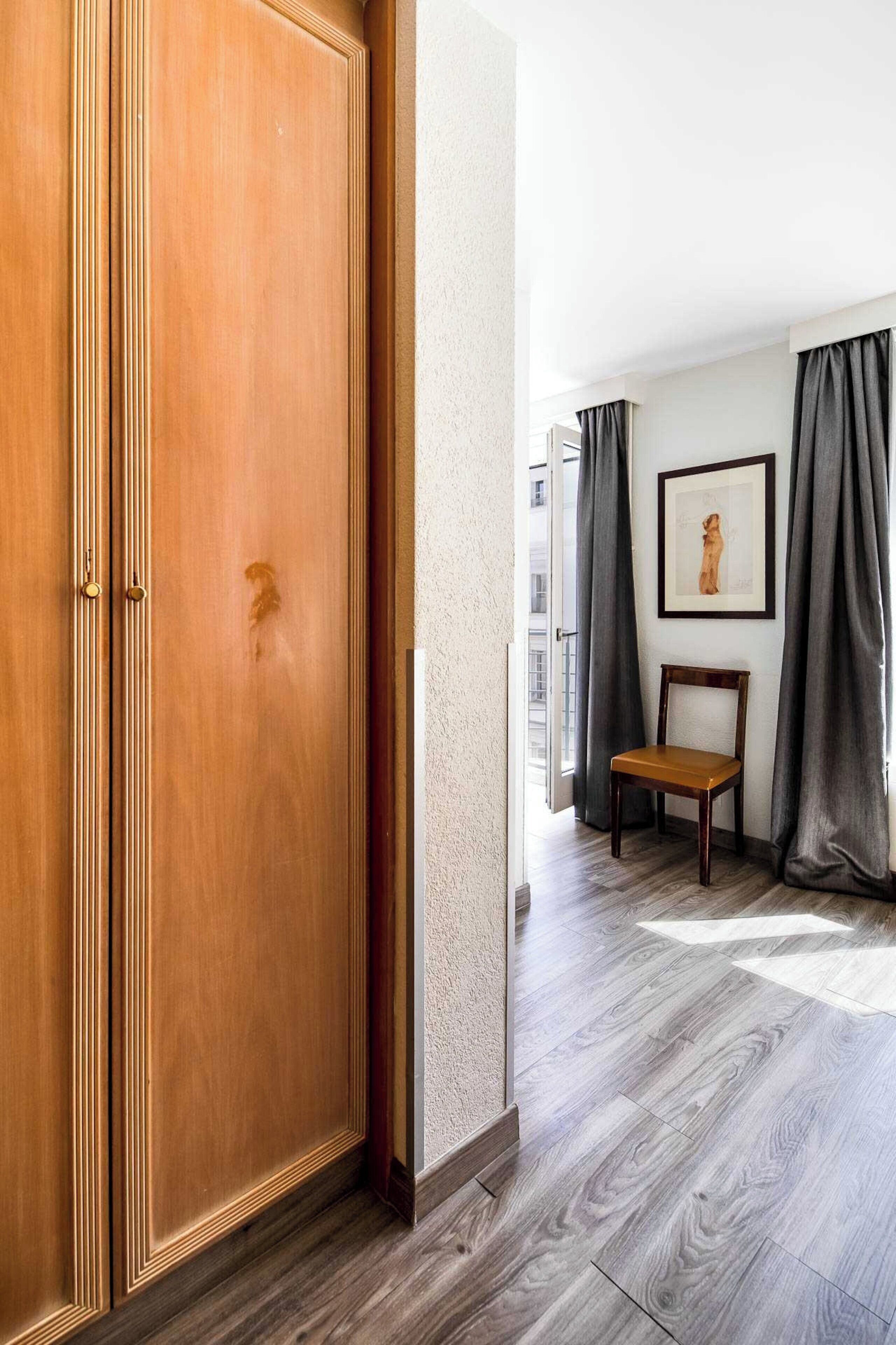 Une pièce avec une armoire en bois, une chaise en bois, un tableau encadré au mur et des rideaux gris.