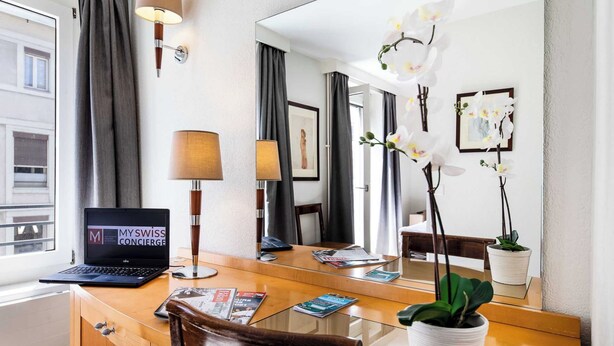 A hotel room with a wooden desk, a laptop, a lamp, a mirror, a potted plant, and a window with curtains. - Charming Apartement- 1br-4p- Geneve (Geneva)