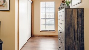 Un pasillo con suelo de madera, una puerta corredera, un ventanal con persianas y un mueble de madera oscuro con cajones.