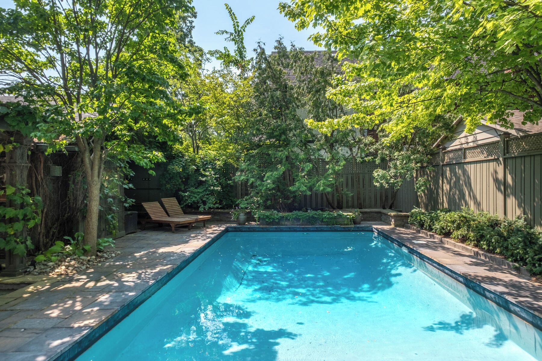 A rectangular swimming pool with a stone border, surrounded by a wooden fence and lush greenery.