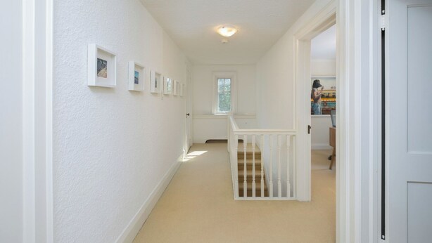 Un couloir aux murs blancs, doté d’un sol recouvert de moquette, d’un escalier avec une rampe blanche et d’une porte donnant sur une autre pièce.