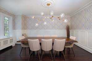 Une salle à manger avec une table en bois, six chaises, un lustre, un papier peint à motifs et une fenêtre donnant sur un beau paysage.