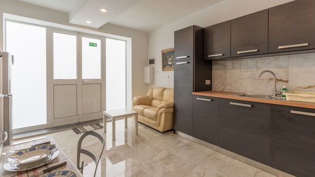 A modern kitchen with dark cabinets, a marble backsplash, and a sink. There is a dining area with a table set for two and a beige sofa. - Maltese Cozy Studio Apartment (SAN PAWL IL-BAĦAR)