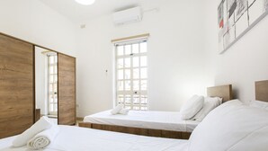 A hotel room with two beds, a wooden wardrobe, a large window, and a ceiling air conditioner.