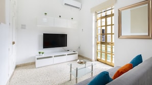 A living room with a flat-screen TV on a white cabinet, a glass coffee table, and a grey sofa with colorful cushions.