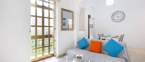 Una sala de estar con una mesa de centro de vidrio, un sofá con cojines de colores, una mesa de comedor con sillas, un reloj de pared y un ventanal con vistas a la vegetación.