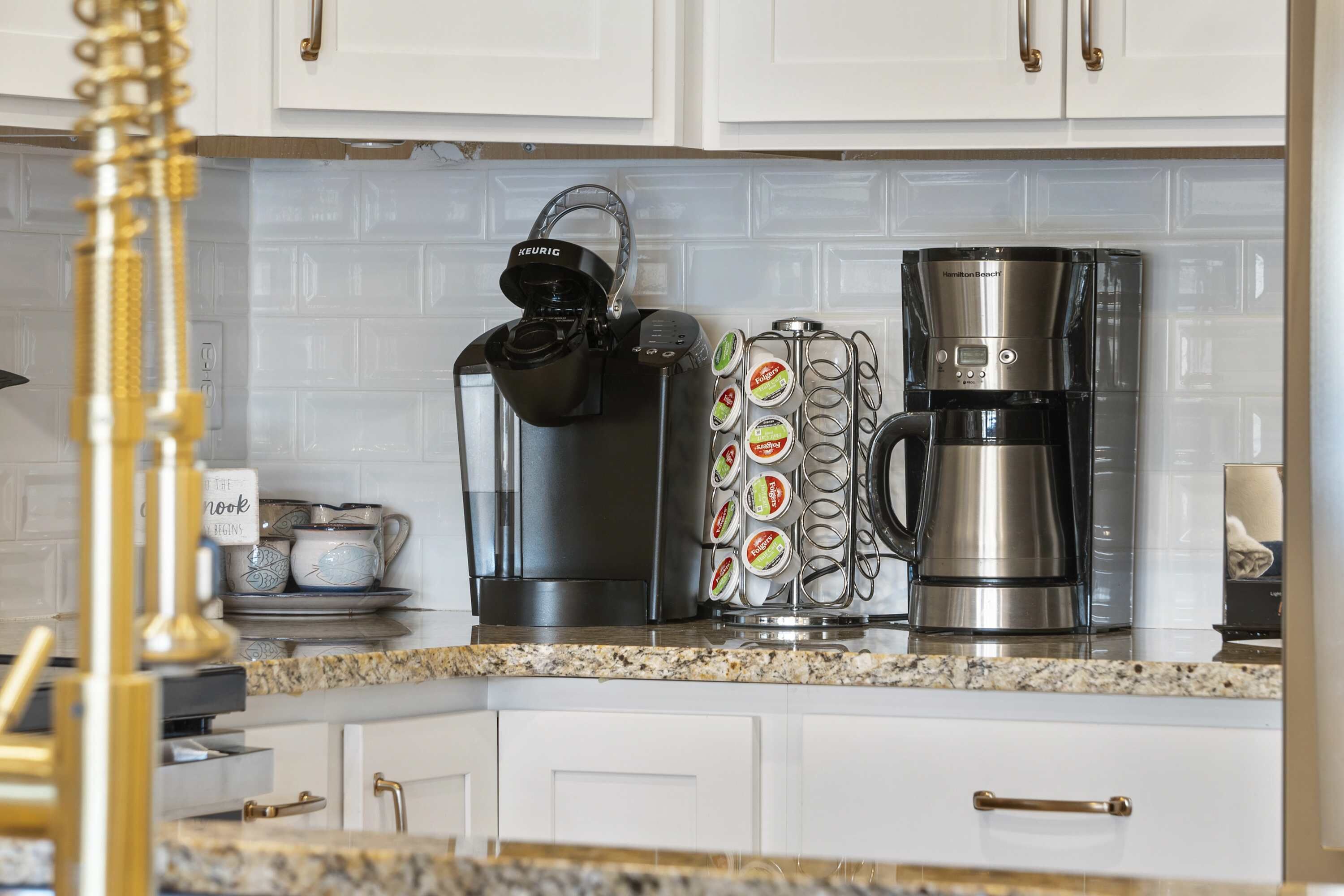 A kitchen with white cabinets, a coffee maker, a kettle, and a mug holder with coffee pods.