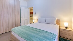A bedroom with a bed, bedside lamps, a wooden wardrobe, and a door leading to a bathroom. - Modern 2-Bedroom Maisonette in Swieqi (Swieqi)