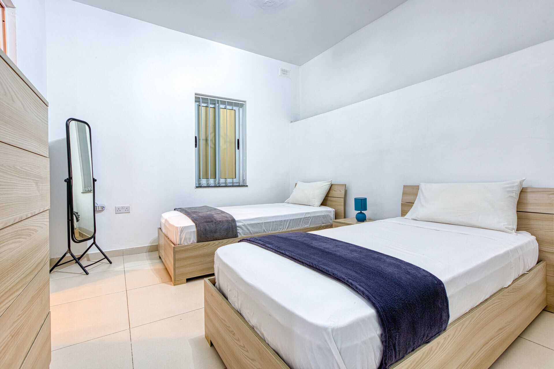 A bedroom with two beds, a window, a mirror, and a bedside table with a lamp.