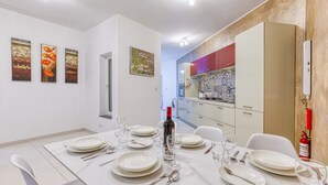 A dining area with a white table set for four, surrounded by white chairs. There are three paintings on the wall, a kitchen with colorful tiles, and a white ceiling with recessed lighting. - Modern 2-Bedroom Maisonette in Swieqi (Swieqi)