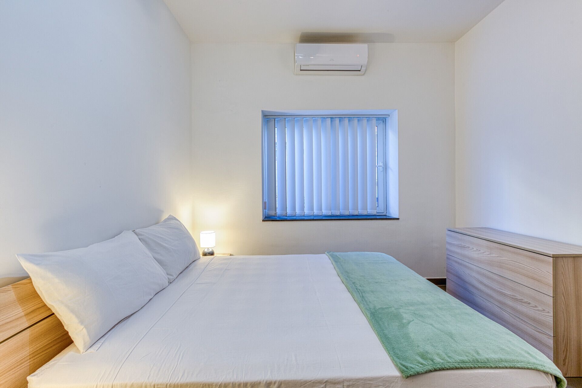 Ein Schlafzimmer mit einem Bett, einem Nachttisch mit Lampe, einem Fenster mit Jalousien und einem Holzkleiderschrank.