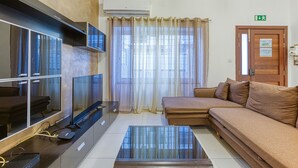 A modern living room with a sofa, a coffee table, and a TV. There is a window with curtains, an air conditioning unit, and a door with an exit sign. - Modern 2-Bedroom Maisonette in Swieqi (Swieqi)