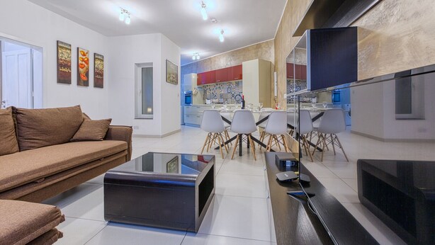 A modern living room with a sofa, a coffee table, and a TV. There is a kitchen in the background with a dining area. - Modern 2-Bedroom Maisonette in Swieqi (Swieqi)