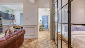 A modern apartment with a wooden herringbone floor, a leather sofa, a bed with a headboard, and a kitchen area with cabinets and a dining table. - Valletta in Style beautiful apartment (IL-BELT VALLETTA)
