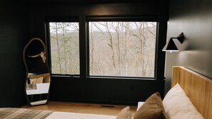 A bedroom with a large window, a bed, a bedside table, and a mirror.
