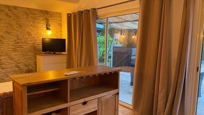 A room with a stone wall, a television, a wooden cabinet, and a sliding glass door leading to an outdoor area. - Tamarind Duo (Arles)