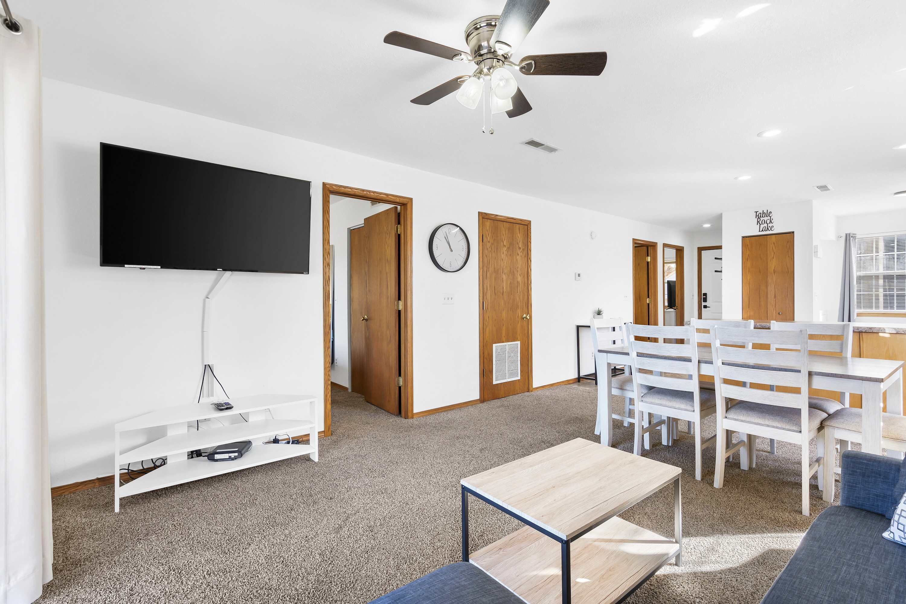 A modern living room with a flat-screen TV, a ceiling fan, a dining area with white furniture, and a clock on the wall.