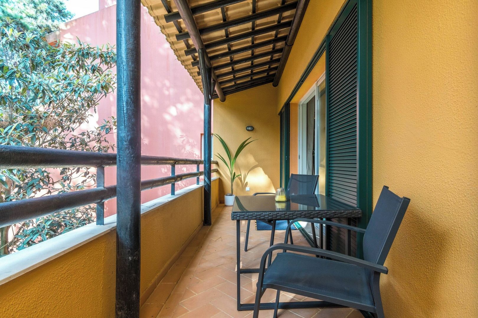 A balcony with a table and chairs, a potted plant, and a view of a red building.