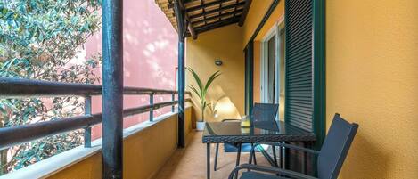 A balcony with a table and chairs, a potted plant, and a view of a red building.