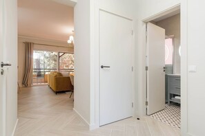 A hallway leading to a living area with a sofa and a dining table, a white door, and a bathroom with a toilet and sink.