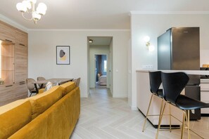 Una cocina moderna con un refrigerador de acero inoxidable, un área de comedor con un sofá amarillo y sillas negras, y un pasillo que conduce a un dormitorio.