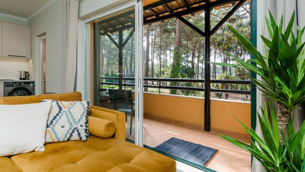 A room with a yellow sofa, a large sliding glass door leading to an outdoor balcony with a view of trees. - Stunning 2 bedroom apartment in Herdade da Aroeira (Almada)