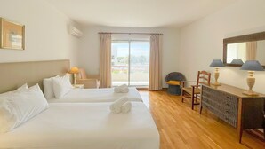 A hotel room with two beds, a desk, a chair, a lamp, and a large window with curtains. - Poolside Bliss T1 Hsrentals Vilamarina (Loulé)