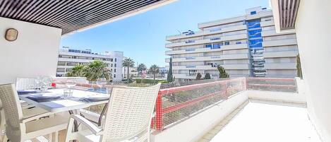 Un balcon avec une table et des chaises blanches, donnant sur un paysage urbain composé d’immeubles modernes et de palmiers.