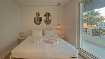A hotel room with a bed, two wall art pieces, a nightstand, a lamp, and a view of palm trees through a window.