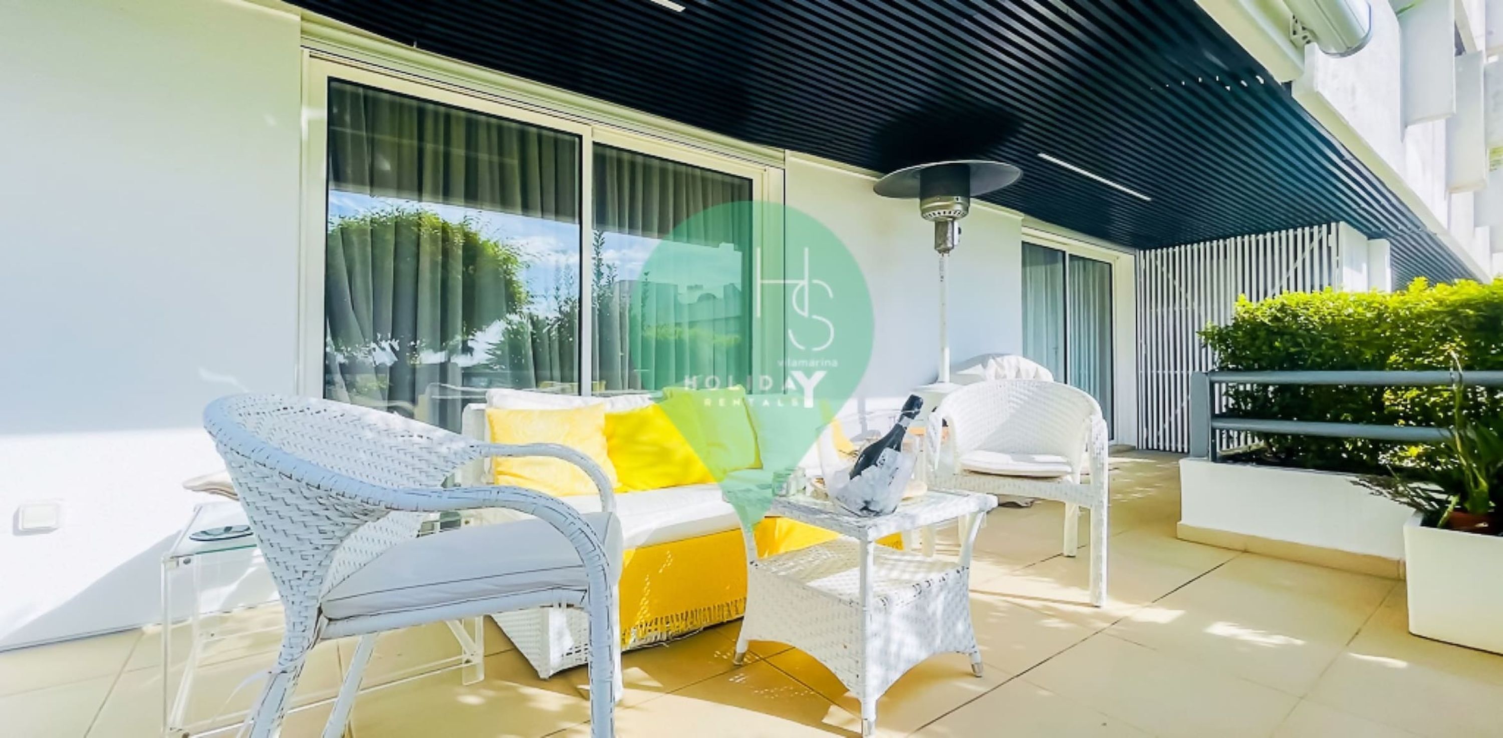 A modern outdoor seating area with wicker chairs and a yellow sofa, a table with a bottle, and a lamp.