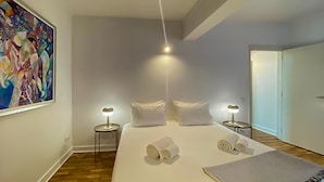 A modern bedroom with a large bed, two bedside tables with lamps, a painting on the wall, and a door leading to another room. - Que Sonho Vista Marina T2 Hsrentals Vilamarina (Loulé)
