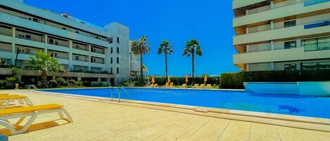 A swimming pool with sun loungers and palm trees, surrounded by modern buildings.