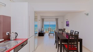 Una cocina moderna con encimera roja, cafetera y lavavajillas. Hay un comedor con mesa y sillas, y vistas al mar a través de grandes ventanales.