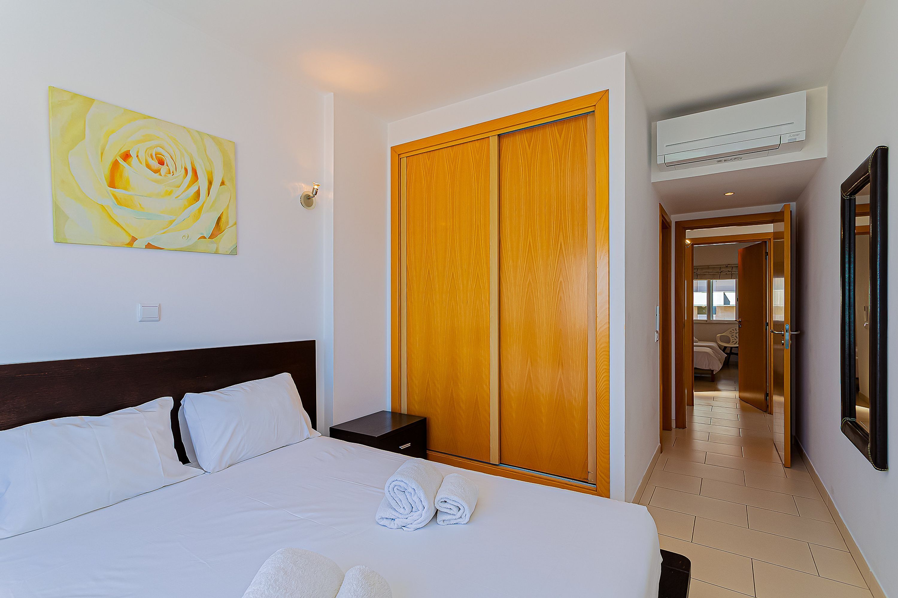 Une chambre d’hôtel avec un grand lit, une armoire en bois, un appareil de climatisation et un tableau représentant une rose accroché au mur.