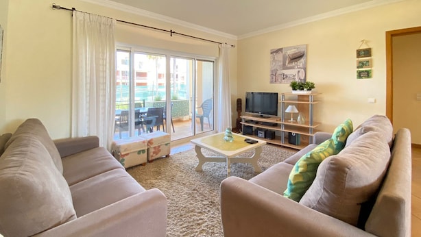 A living room with two sofas, a coffee table, a TV, and a sliding glass door leading to an outdoor area with chairs. - Marina Sol T3 Hsrentals Vilamoura (Loulé)