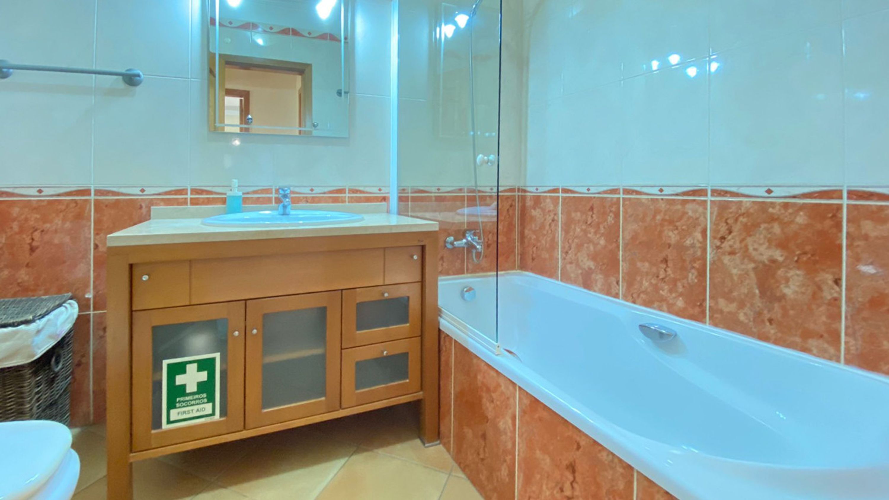 Un baño con bañera, lavamanos y espejo. Hay un cartel en el mueble que indica botiquín de primeros auxilios.