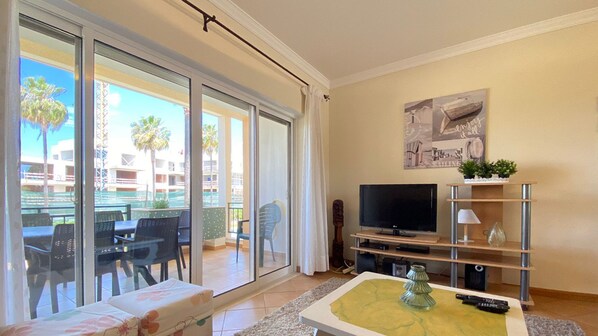 Una sala de estar con televisión, sofá, mesa de centro y puerta corrediza de vidrio que da a un área exterior con sillas y una mesa.