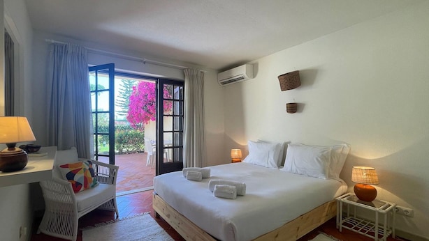 A hotel room with a bed, bedside lamps, a desk with a lamp, a chair, and a view of a garden through an open door.