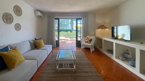 A living room with a white sofa, a coffee table, a TV on a shelf, and a view through a glass door to an outdoor area.