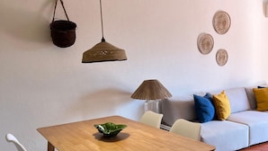 A dining area with a wooden table, white chairs, a hanging basket, a woven lamp, and a sofa with cushions.
